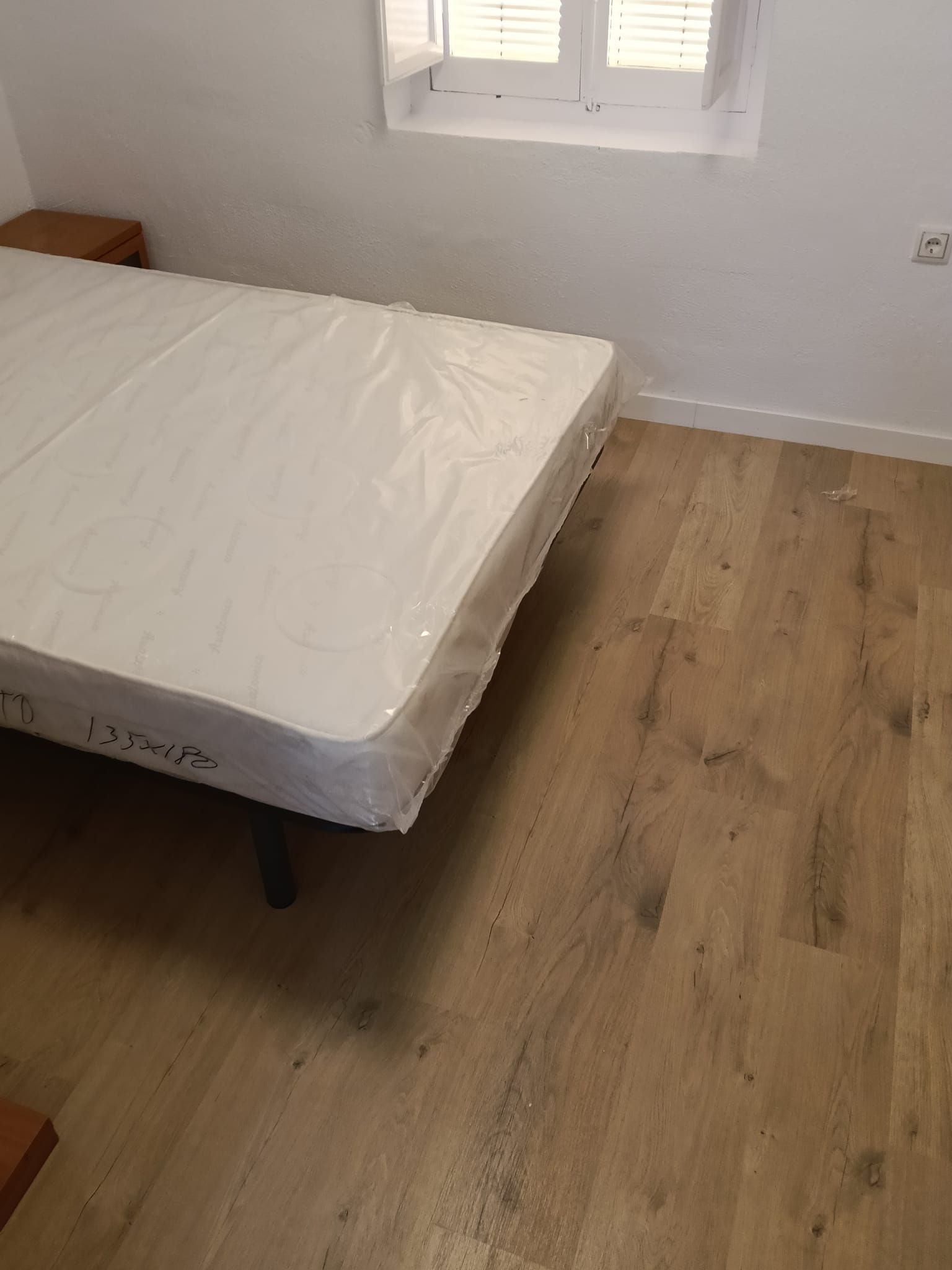 Cama sin hacer con colchón blanco en una habitación pequeña con suelo de madera clara y una ventana.