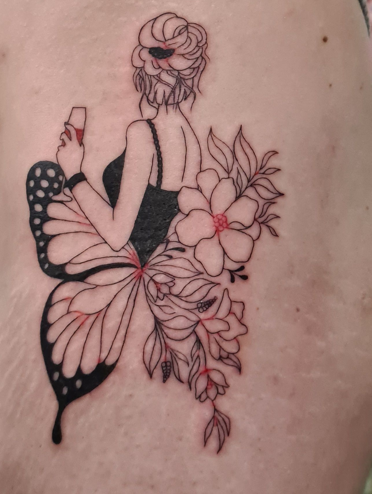 Tatouage d'une femme aux ailes de papillon, tenant un verre. Des fleurs ornent ses cheveux.