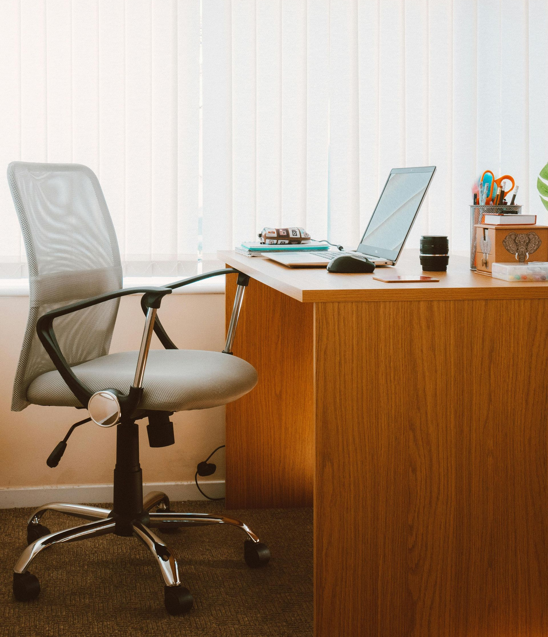 Un bureau avec un ordinateur portable, une chaise de bureau et divers objets posés sur le bureau, devant une fenêtre.