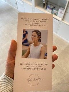 Eine Person hält eine Broschüre mit einem Foto einer meditierenden Frau, der Website-Adresse und den Kontaktinformationen in der Hand.