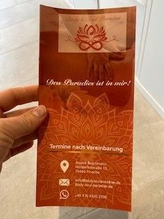 Eine Broschüre wird in der Hand gehalten. Sie ist orangefarben und trägt ein Lotus-Logo sowie Werbung für ein Spa-Angebot. Kontaktdaten sind angegeben.