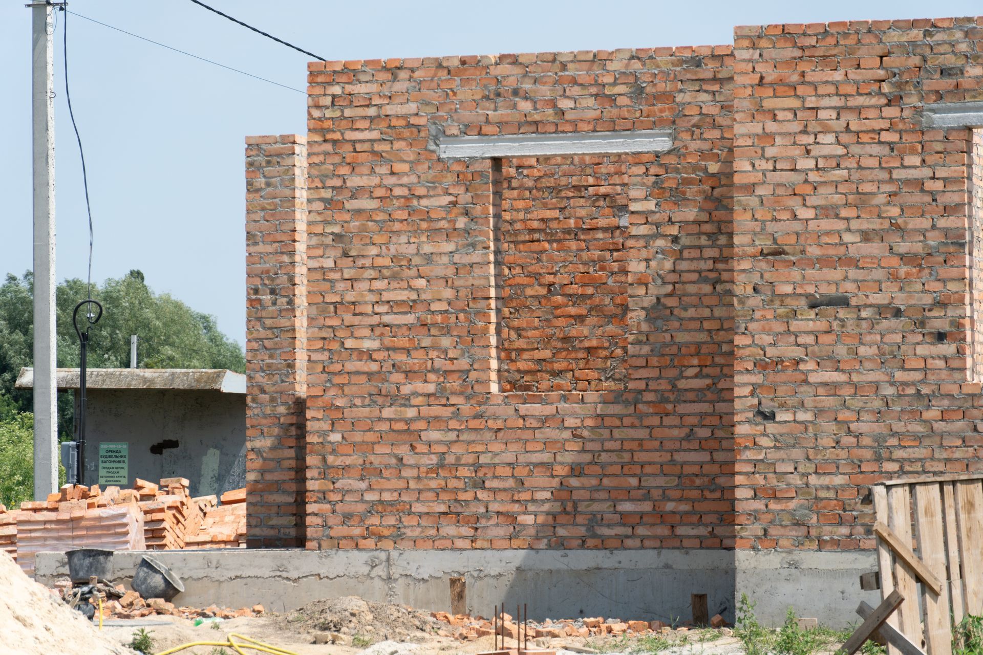 Mur de briques en construction avec ouverture pour fenêtre, briques empilées à proximité, environnement extérieur.