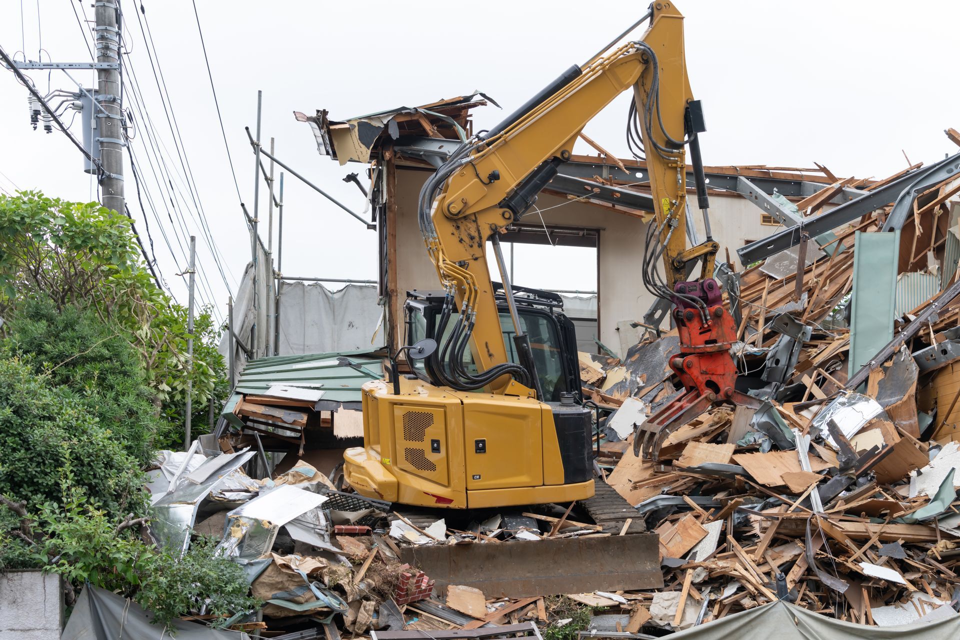 Une pelleteuse jaune démolit une maison, des débris sont éparpillés autour ; ciel couvert.