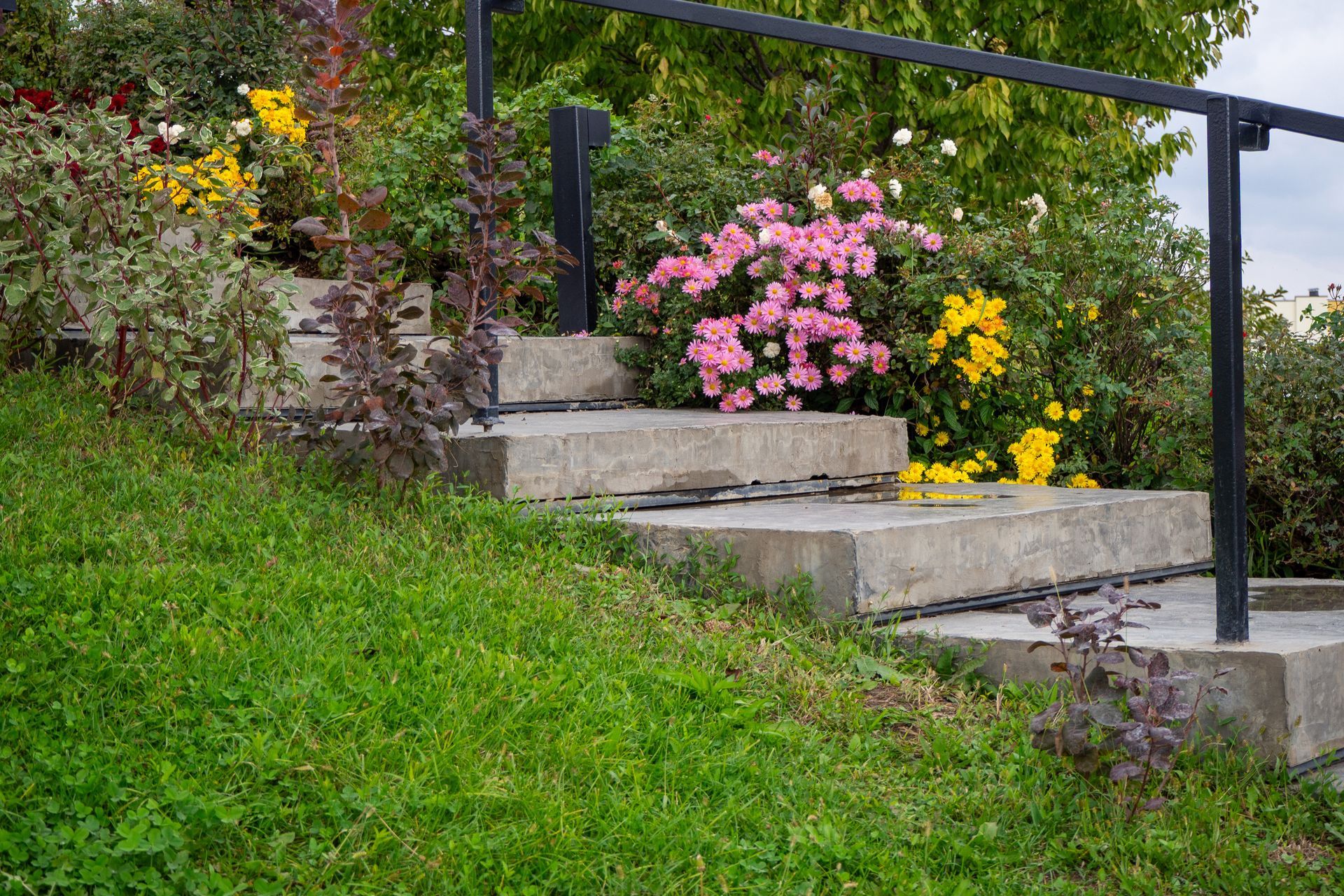 Un escalier en béton avec une rampe noire monte une colline herbeuse, entourée de fleurs colorées.