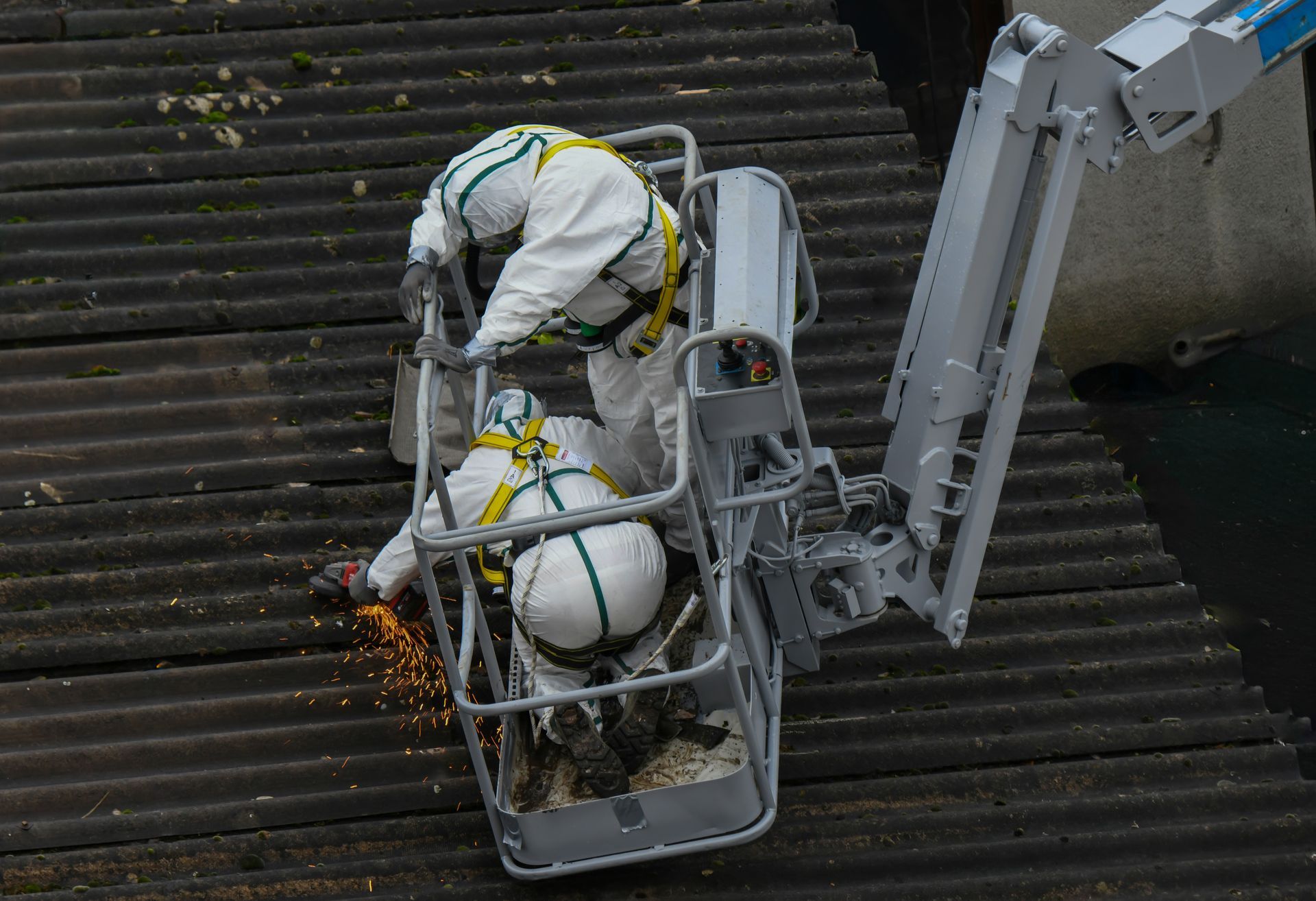 Deux ouvriers en combinaisons de protection sur une nacelle élévatrice découpent des matériaux de toiture, des étincelles jaillissent.