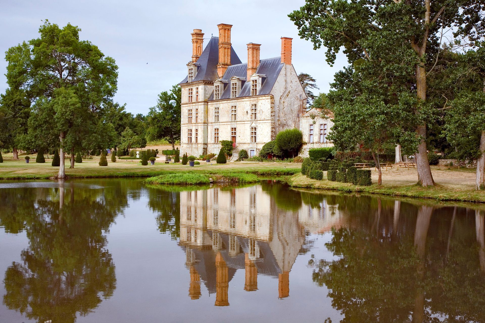 Château de la Motte-Glain se reflétant dans un étang ; bâtiment en pierre, toit orange, arbres et ciel.