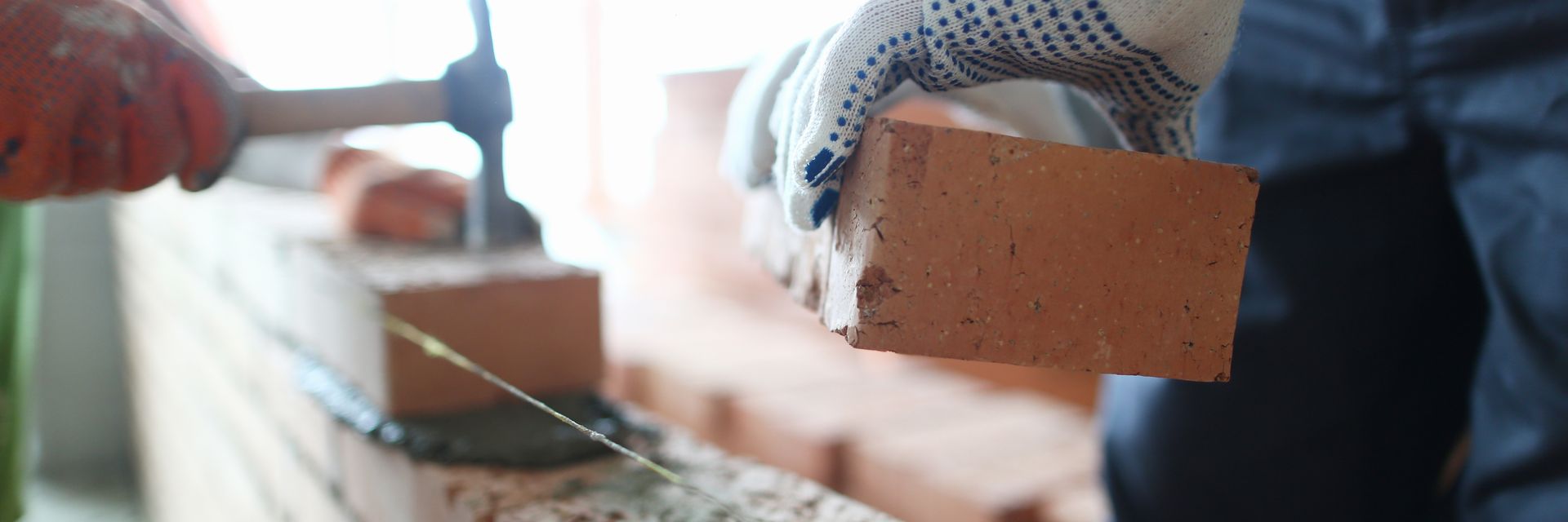 Une personne portant des gants pose une brique sur un mur en construction. Une autre personne utilise un marteau.