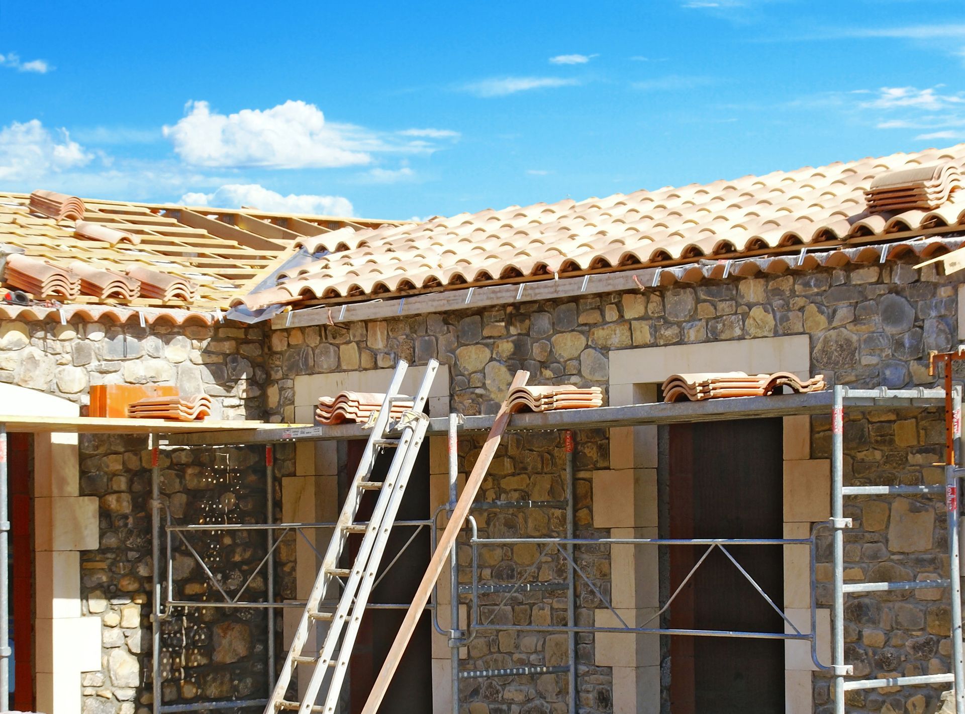 Bâtiment en construction avec des murs en pierre, un toit de tuiles, des échafaudages et une échelle sous un ciel bleu.