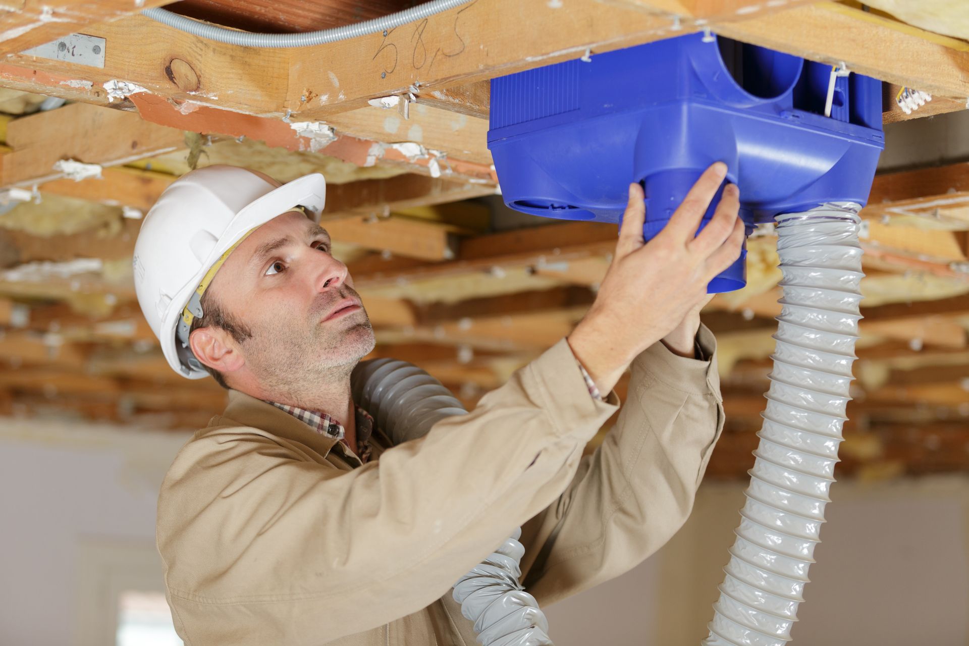 Un homme portant un casque de chantier installe un système de ventilation bleu au plafond.