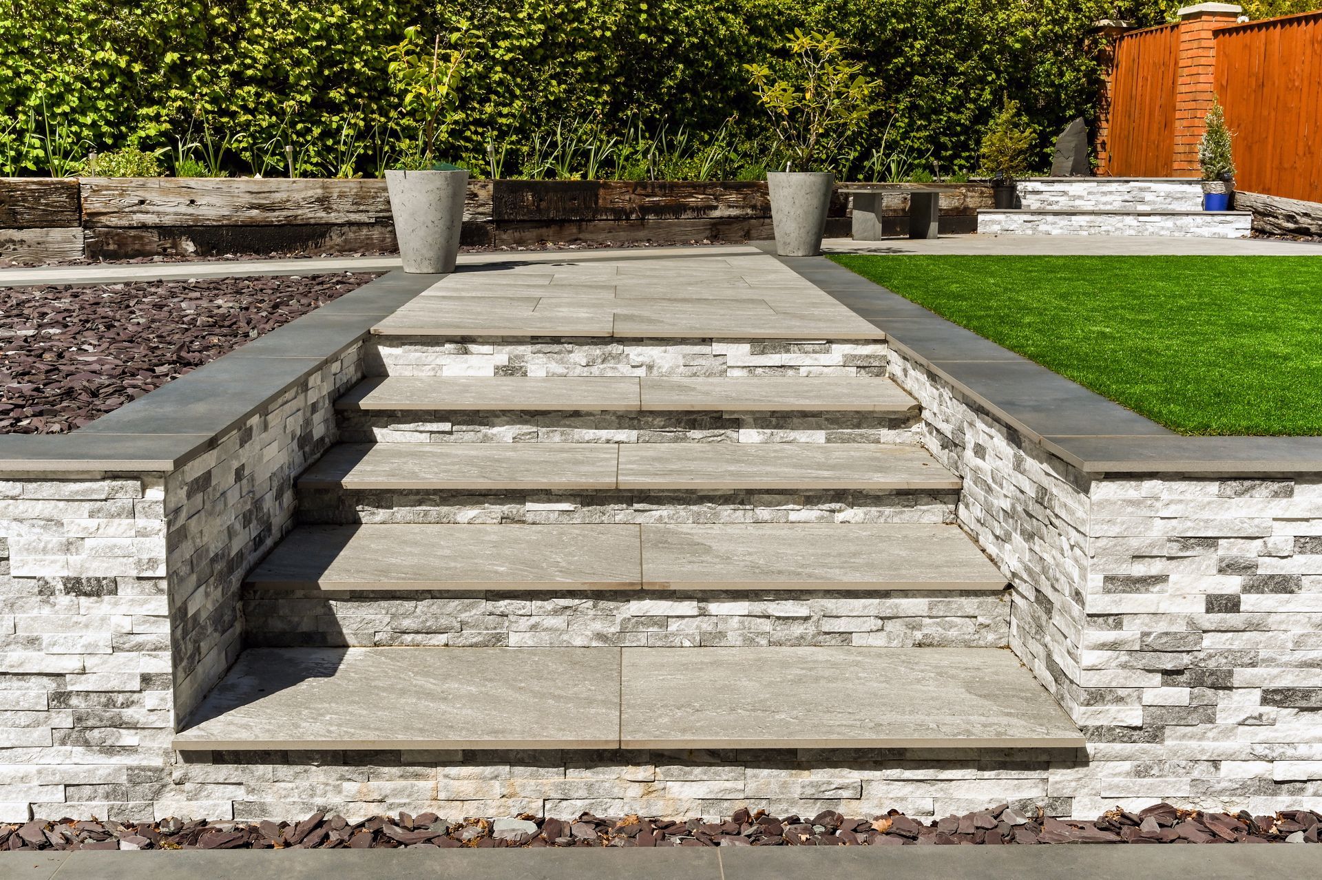 Des marches en pierre mènent à une terrasse ornée de murets et de jardinières. Pelouse verte en arrière-plan.