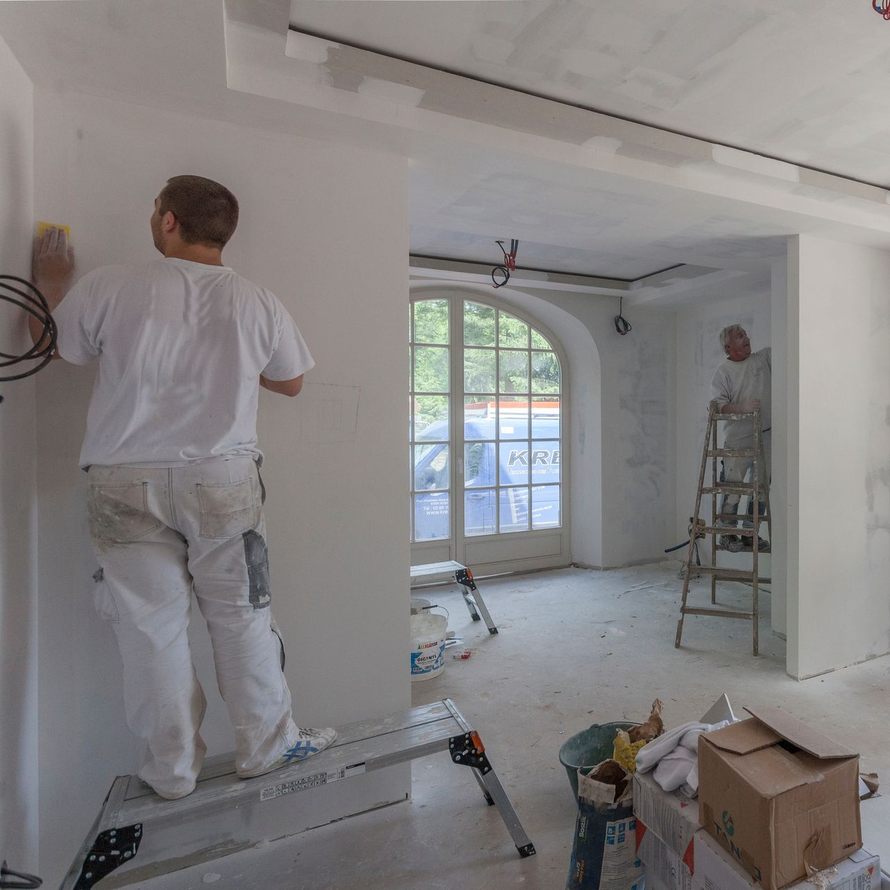 Deux personnes peignent une pièce : l'une sur un escabeau, l'autre debout sur une estrade, toutes deux vêtues de blanc.