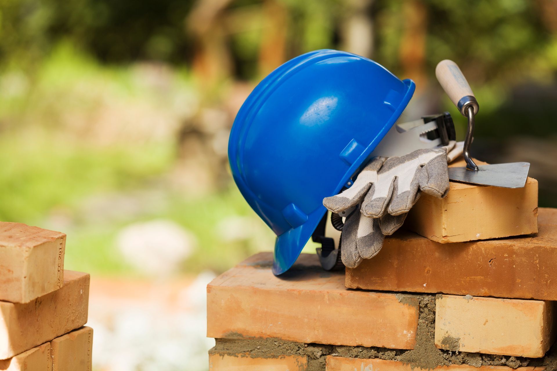 Casque de chantier bleu, gants de travail et truelle posés sur des briques, sur un fond vert flou.