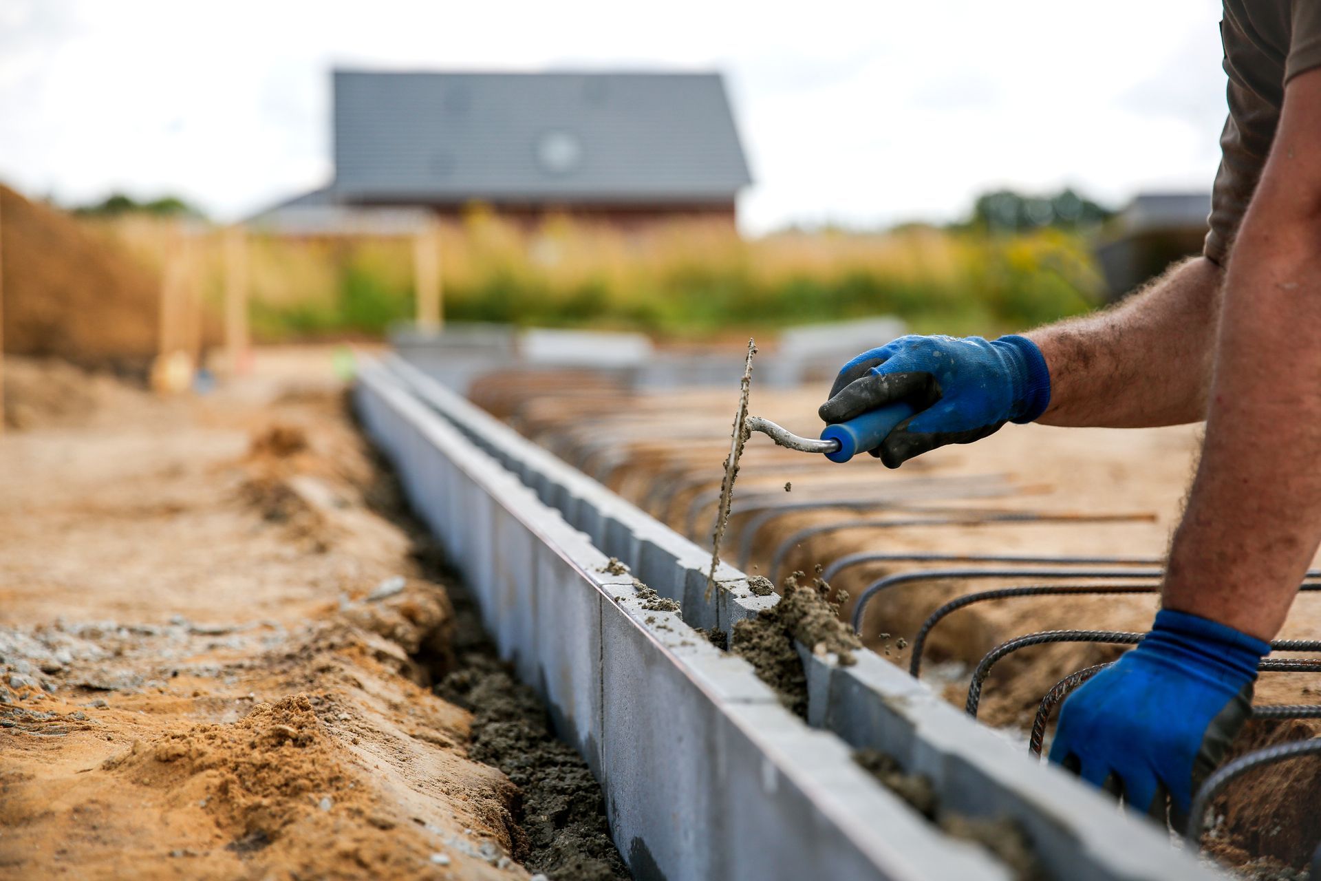 Un ouvrier du bâtiment, portant des gants bleus, étale du ciment à la truelle, en extérieur.