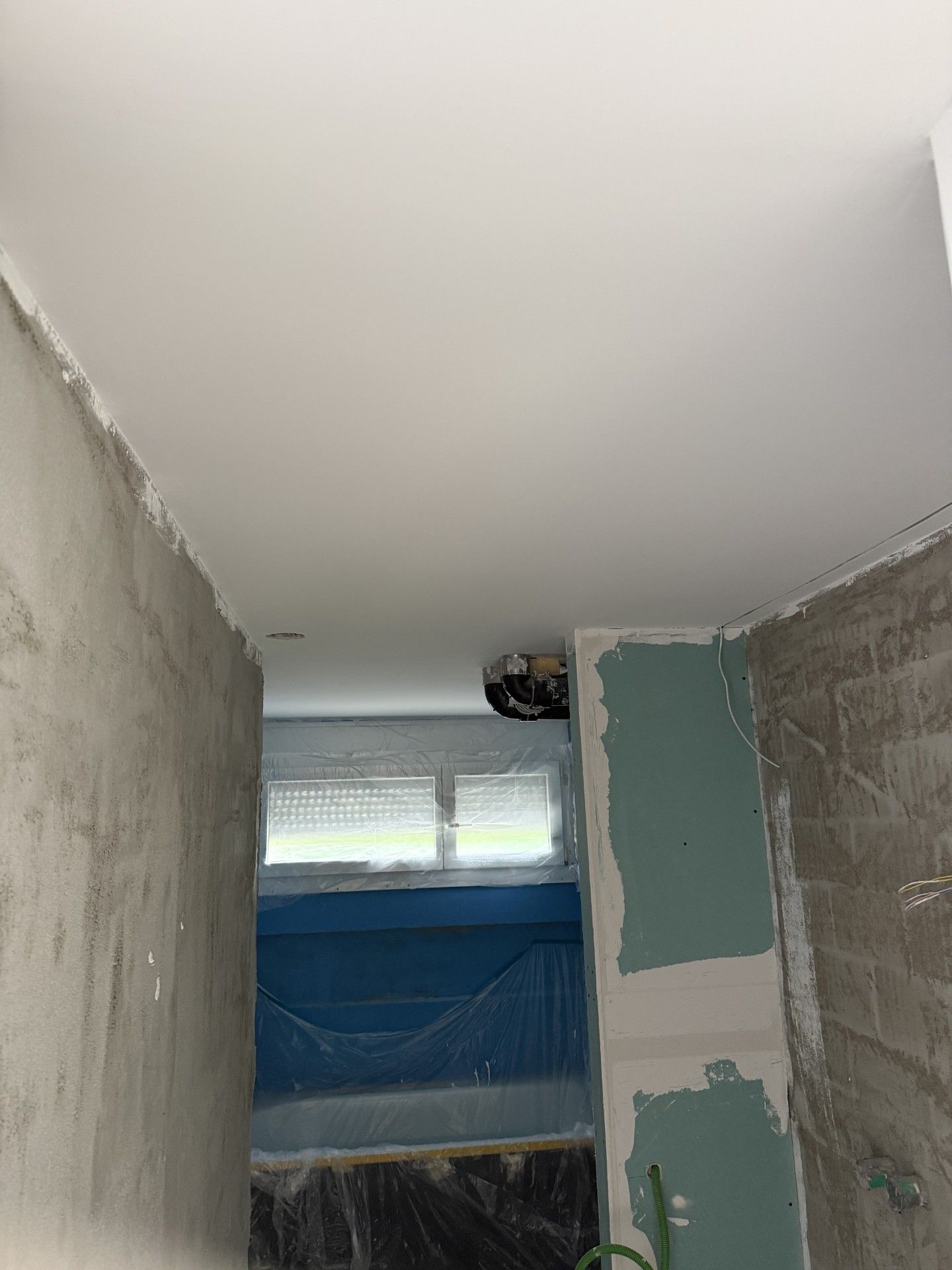 Unfertiges Zimmer mit kahlen Wänden, blau verklebtem Fenster und Ausbesserungsarbeiten an der Decke.