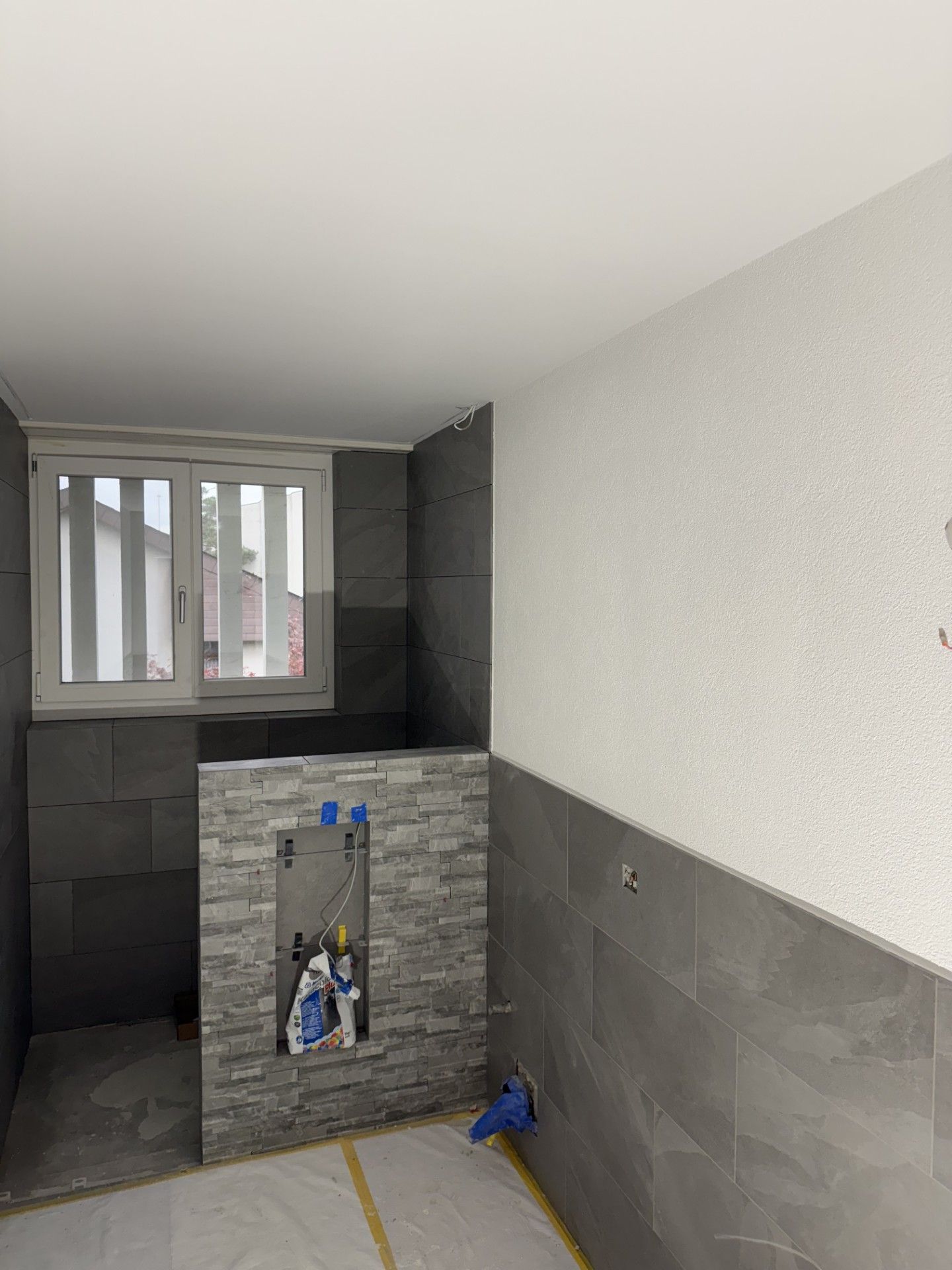 Kleiner, gefliester Hauswirtschaftsraum mit Fenster, grauen Wänden und freiliegenden Rohrleitungen in einem halbfertigen Spülbeckenbereich.