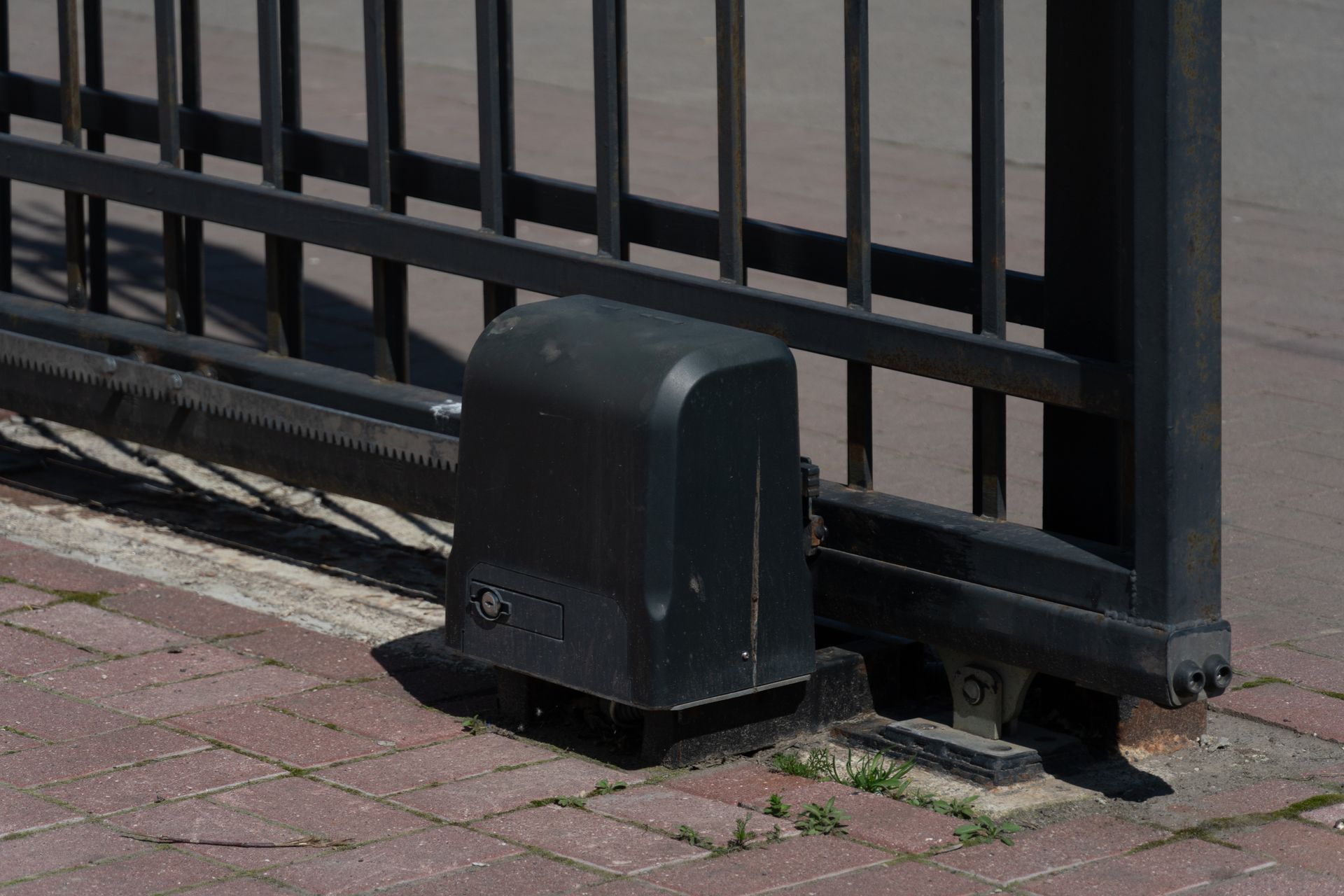 Ouvre-portail automatique noir sur trottoir en briques, fixé à un portail métallique.