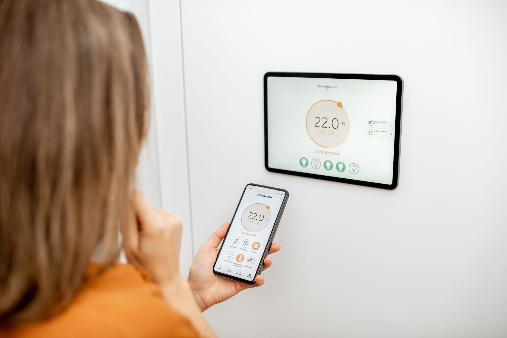 Une femme utilise son smartphone pour contrôler un thermostat intelligent mural, affichant la température.