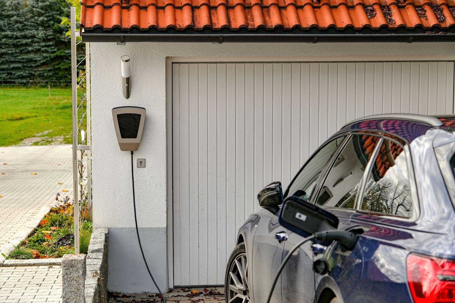 Voiture bleue en charge près d'un garage avec une borne de recharge fixée sur un mur blanc.