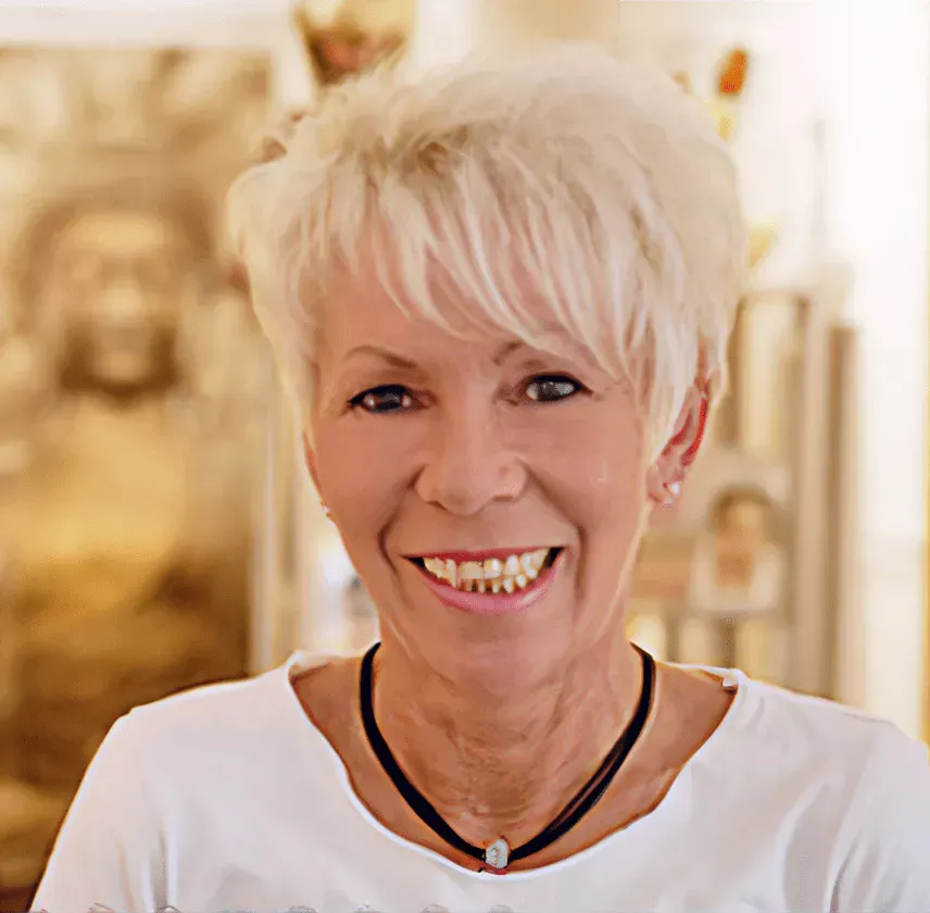 Eine lächelnde Frau mit kurzen blonden Haaren in einem weißen Oberteil steht in einem Innenraum vor einem leicht verschwommenen Hintergrund.