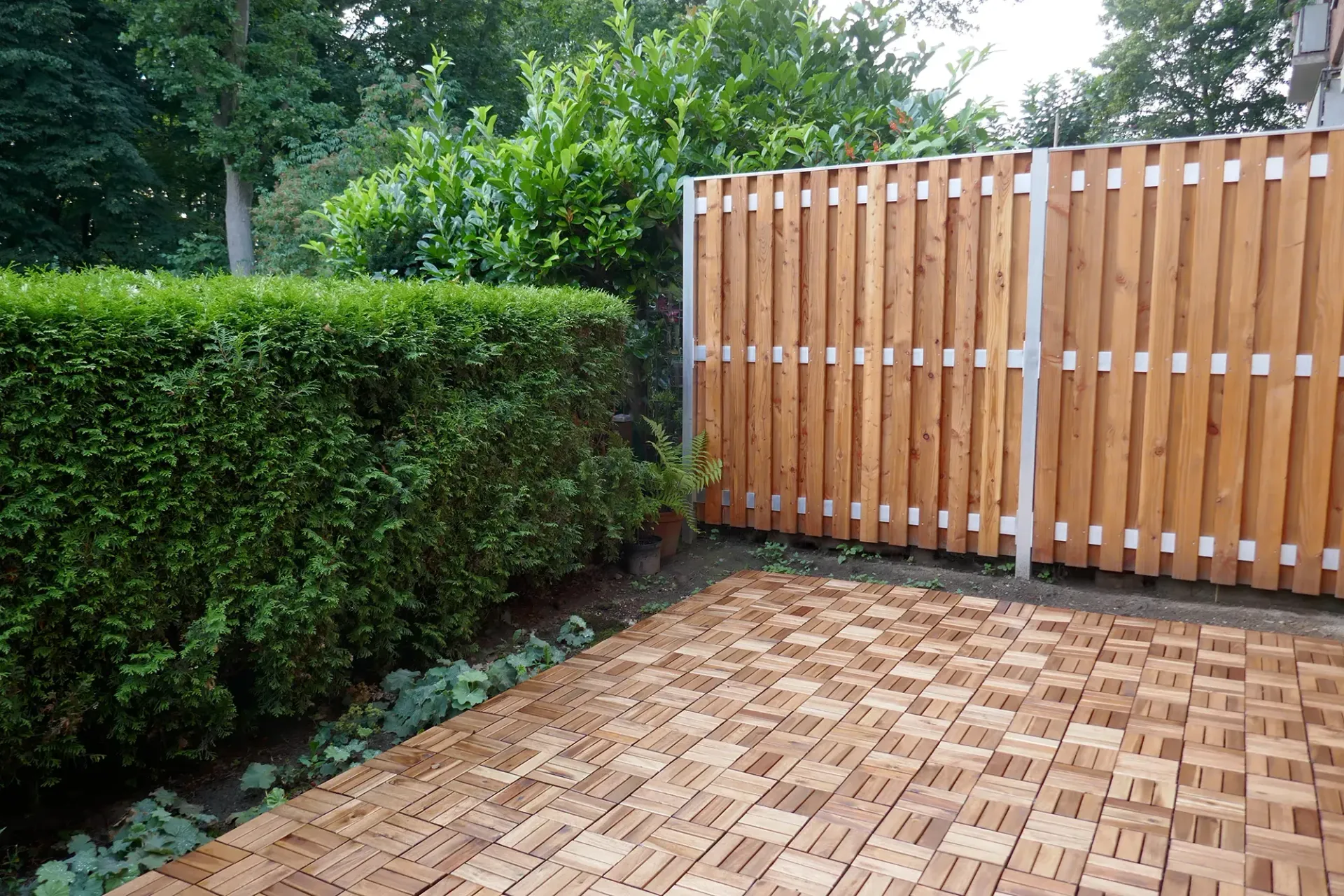 Ein Holzzaun umgibt eine gemauerte Terrasse mit Bäumen im Hintergrund.
