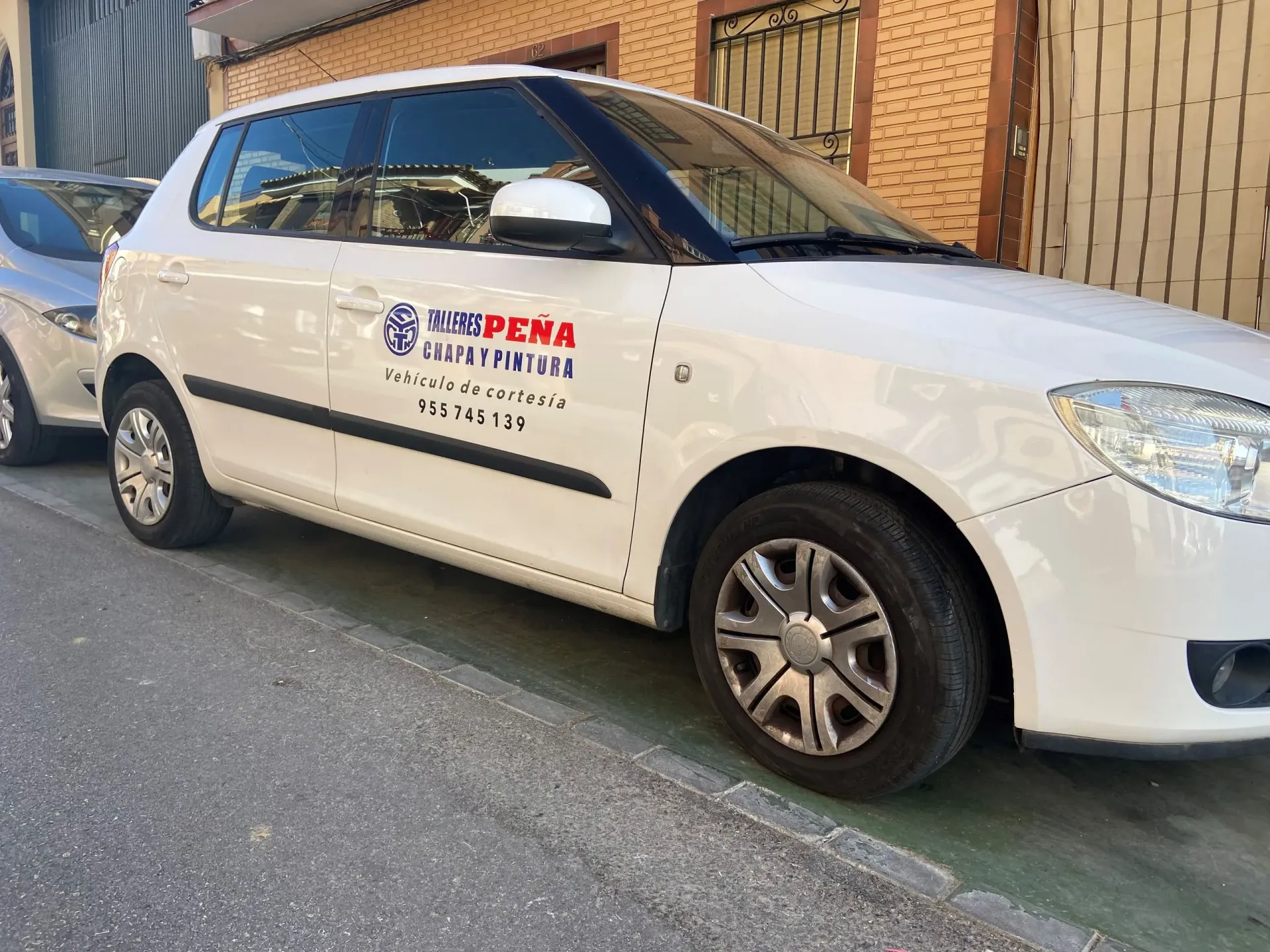 Sedán blanco con el logotipo de la empresa estacionado junto a una acera y la pared de un edificio.