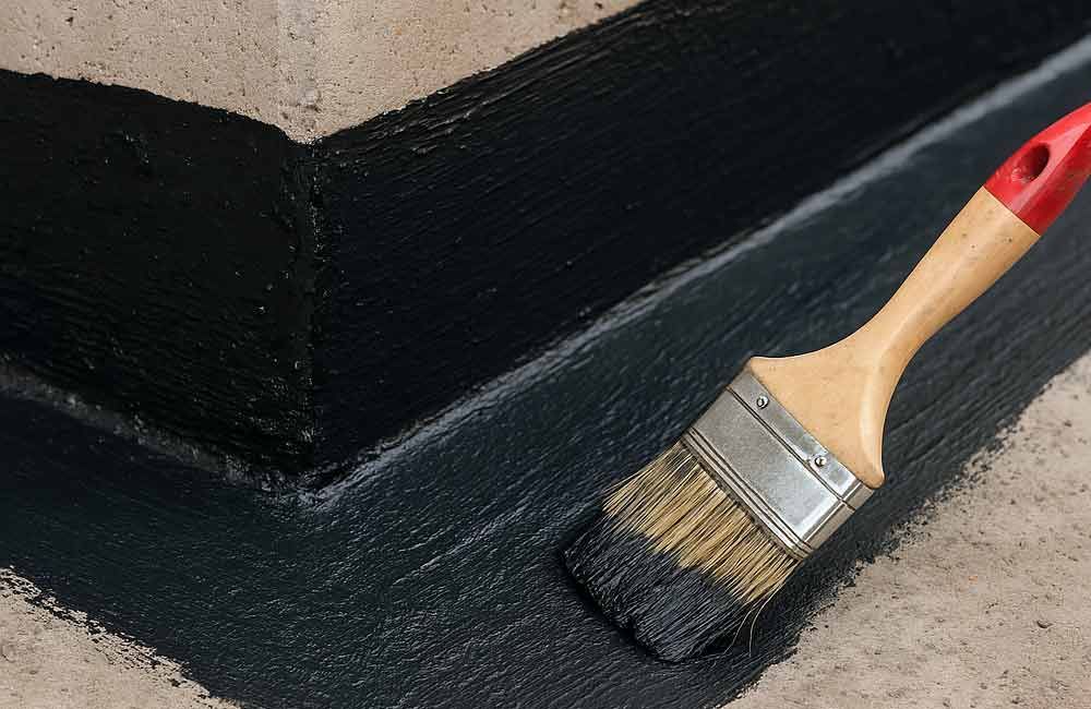 Mit einem Pinsel wird schwarzes Dichtmittel auf eine schräge Betonfläche aufgetragen.