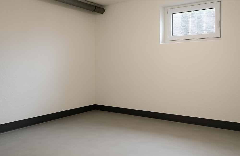 Ein leerer, hellfarbiger Raum mit einem kleinen Fenster, dunklen Fußleisten und einem Rohr an der Decke.