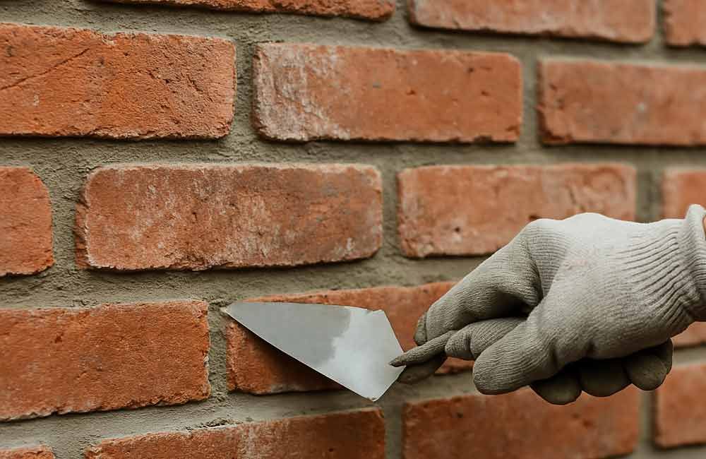 Eine behandschuhte Hand benutzt eine Kelle, um Mörtel zwischen rote Ziegelsteine ​​in einer Mauer aufzutragen.