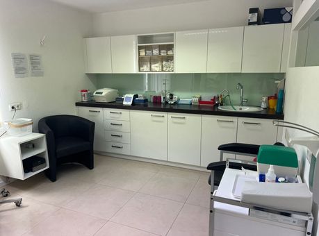 Behandlungsraum mit weißen Schränken, Geräten auf der Arbeitsfläche, einem schwarzen Stuhl und einem Behandlungswagen
