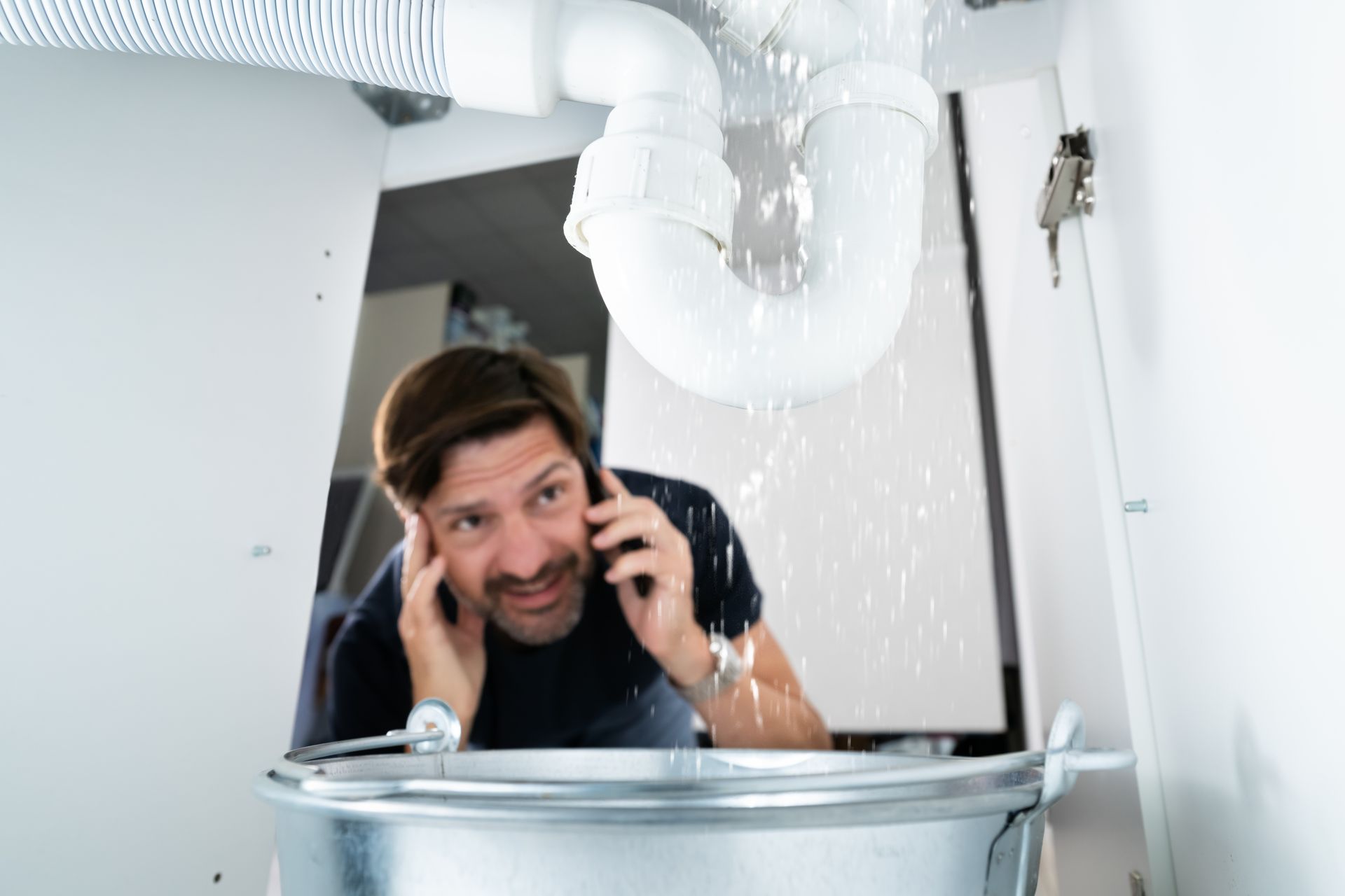 Une personne au téléphone, face à une fuite d'eau en dessous d'un évier.