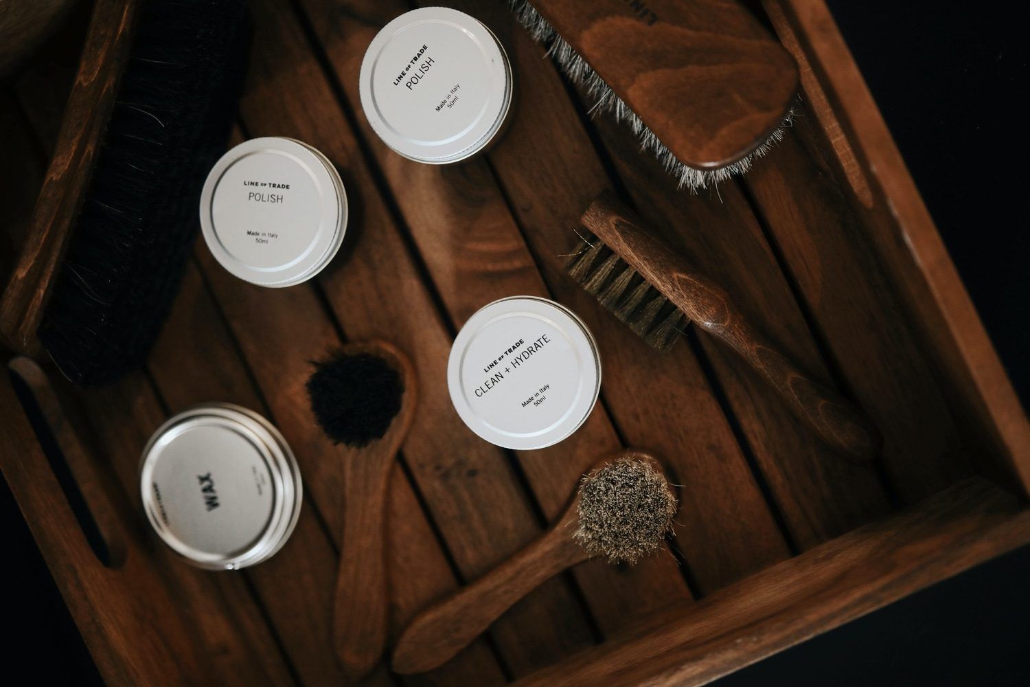 Quatre pots de cosmétiques blancs et des pinceaux disposés dans une boîte en bois sur une surface sombre.