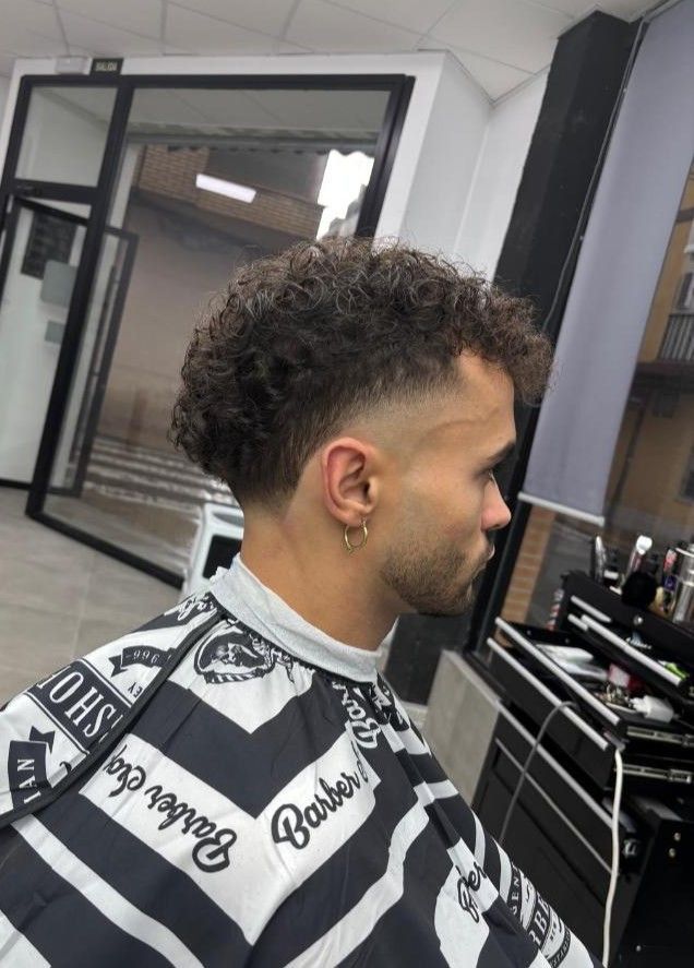 Hombre con cabello rizado y corte de pelo descolorido en una barbería, vistiendo una capa de barbero.