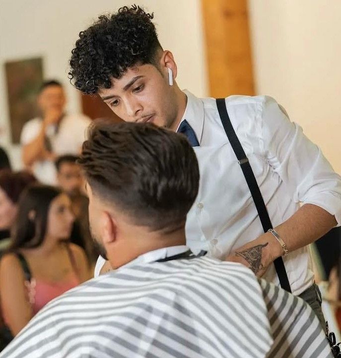 Barbero dando un corte de pelo en el interior; hombre con camisa blanca y tirantes, cliente con capa de rayas.