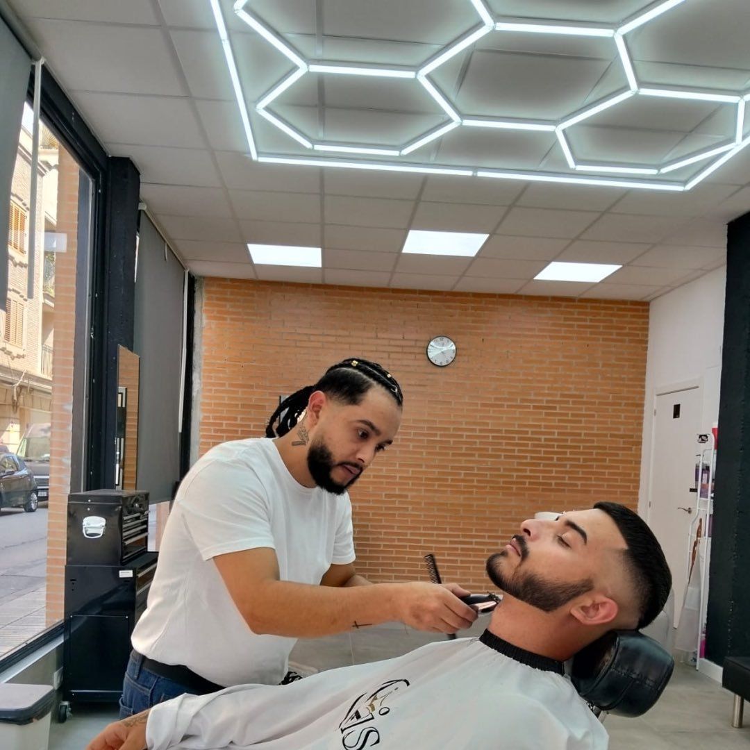 Barbero recortando la barba de un cliente en una barbería con luces de techo en forma de panal.