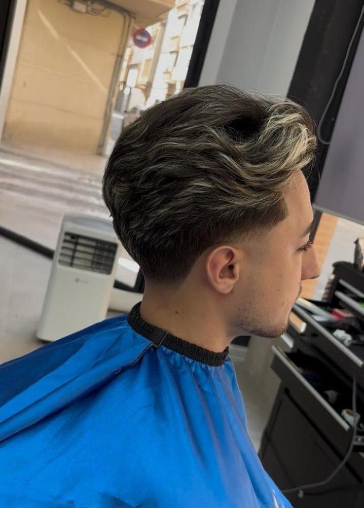 Hombre con peinado ondulado y laterales descoloridos, vistiendo una capa de barbero azul, en un salón.