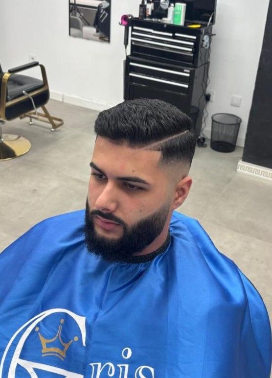 Hombre con un corte de pelo fresco, desvanecido con barba, en una barbería, vistiendo una capa azul.