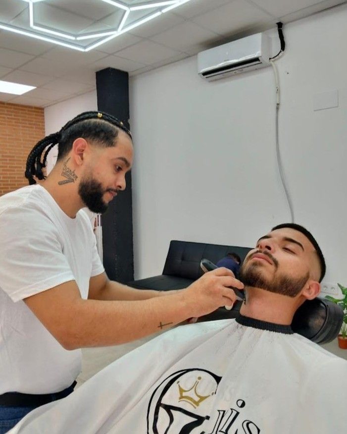 Un barbero le recorta la barba a un cliente en una barbería. El barbero tiene rastas y un tatuaje en el cuello.