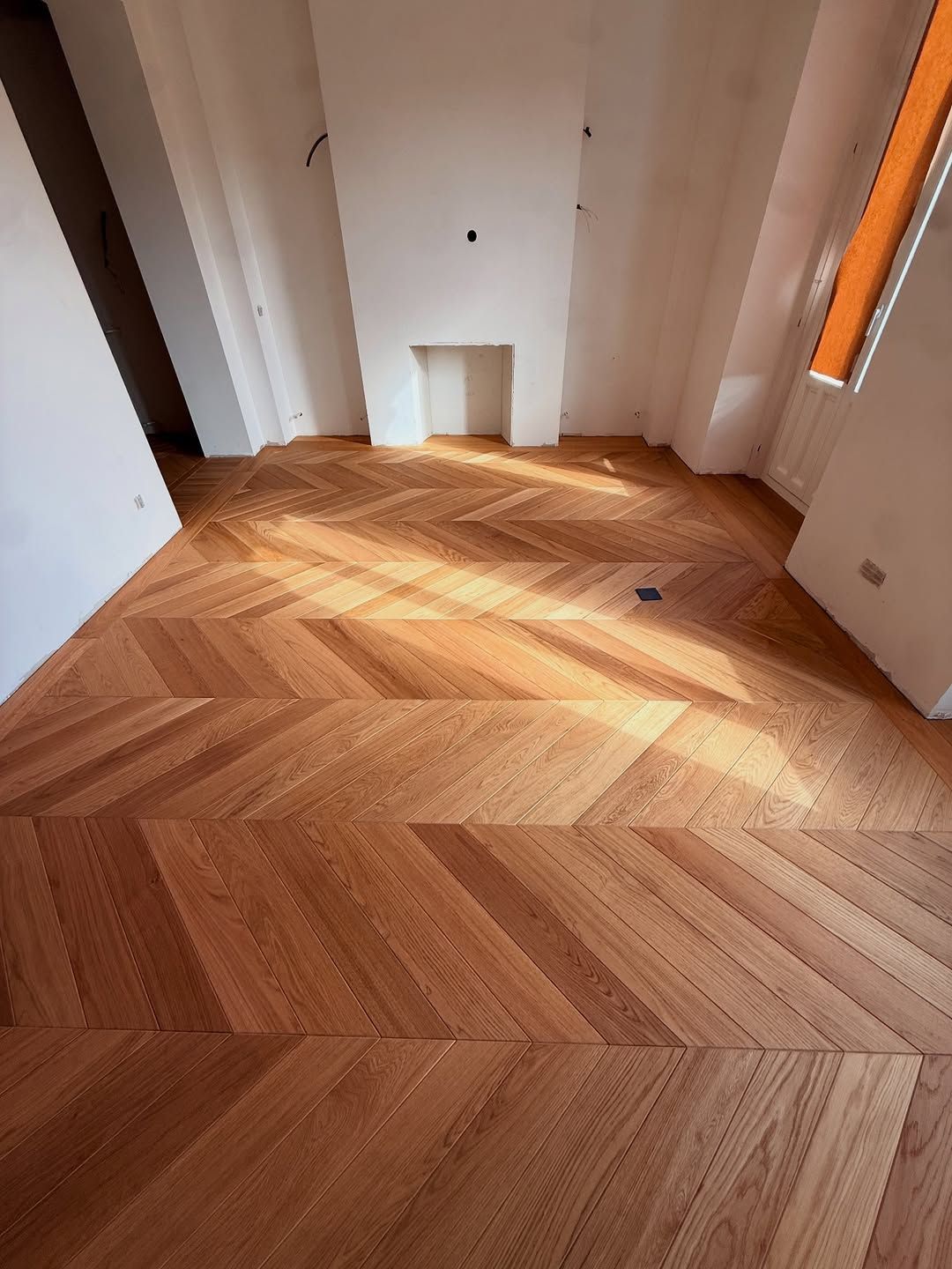 Una habitación con piso de madera recién instalado, de color claro, con diseño de chevrones y paredes blancas.