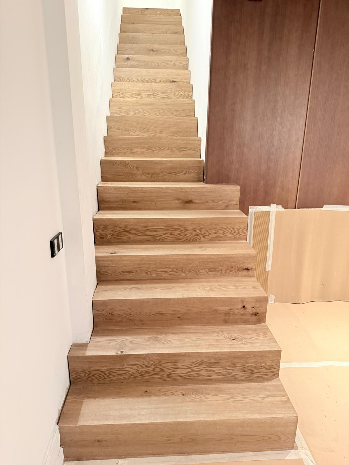 Una escalera recta de madera clara en un espacio interior moderno con paredes blancas.