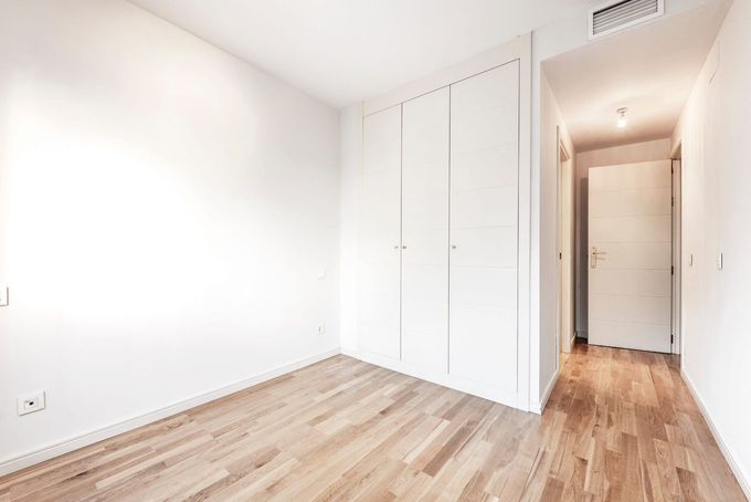 Habitación blanca vacía con suelos de madera, armario empotrado y pasillo que conduce a una puerta.