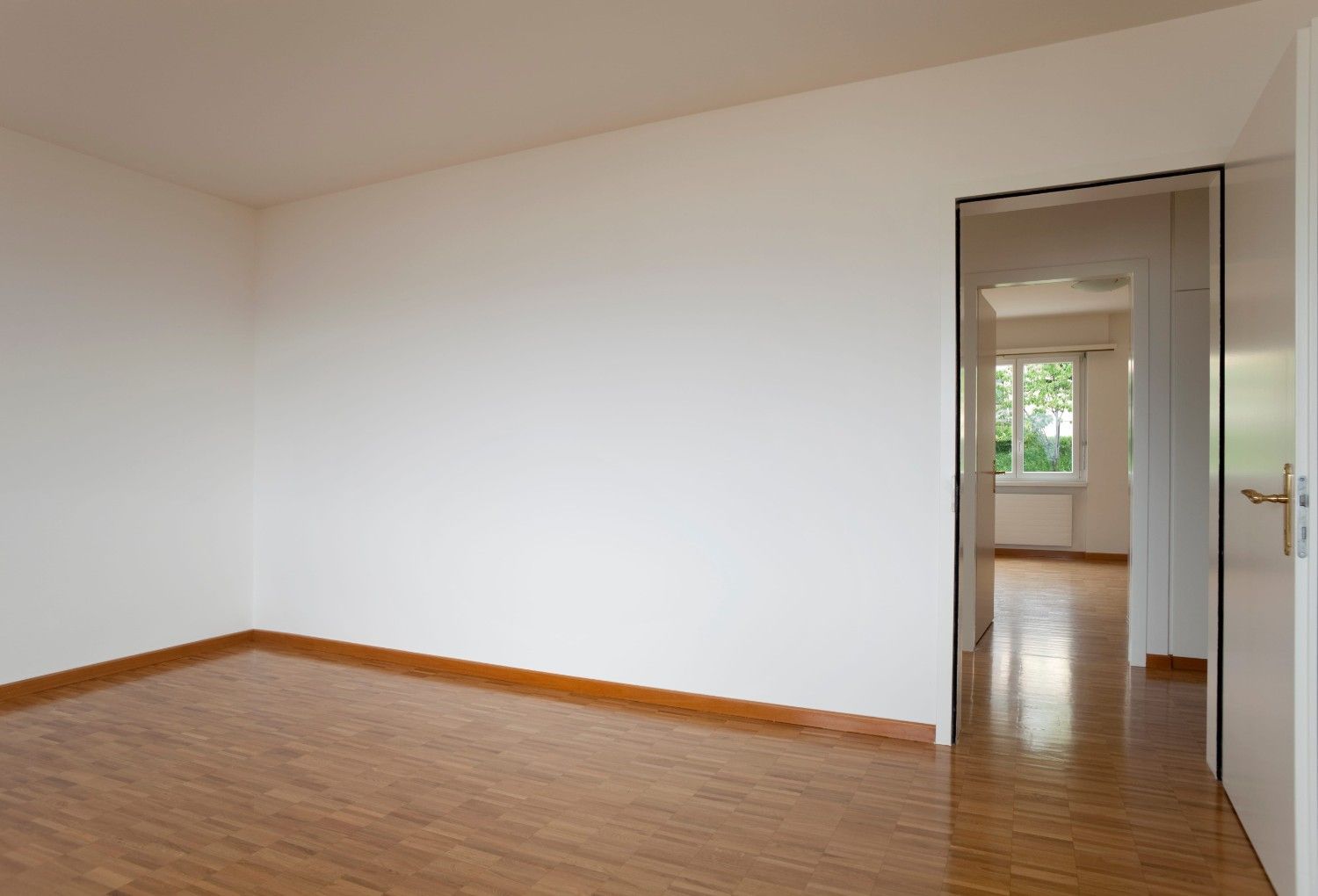 Habitación blanca vacía con suelo de madera y una puerta abierta que da a una habitación contigua luminosa.