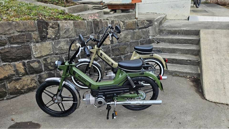 Zwei Oldtimer-Mopeds, eines grün und eines beige, parkten auf einer Betonfläche an einer Steinmauer.