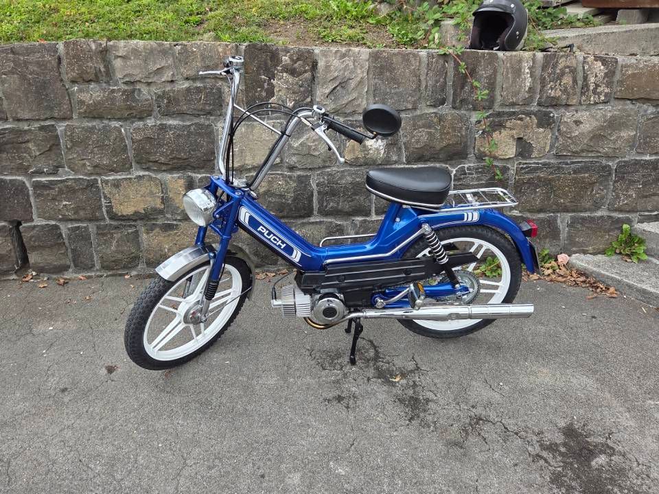 Ein blaues Moped mit Weißwandreifen steht auf Asphalt an einer Steinmauer. Darüber liegt ein schwarzer Helm.