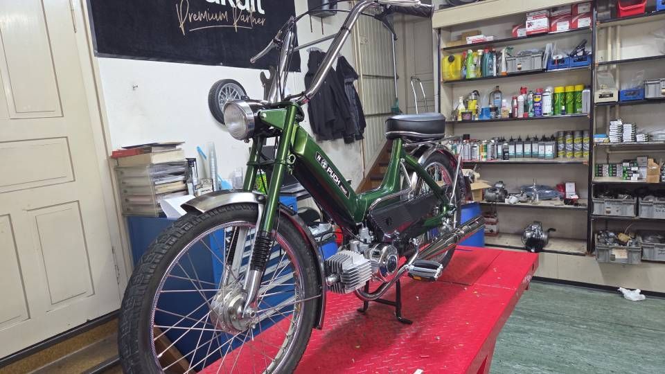 Ein grünes Moped steht auf einem roten Tisch in einer Werkstatt; im Hintergrund sind Werkzeuge und Regale zu sehen.