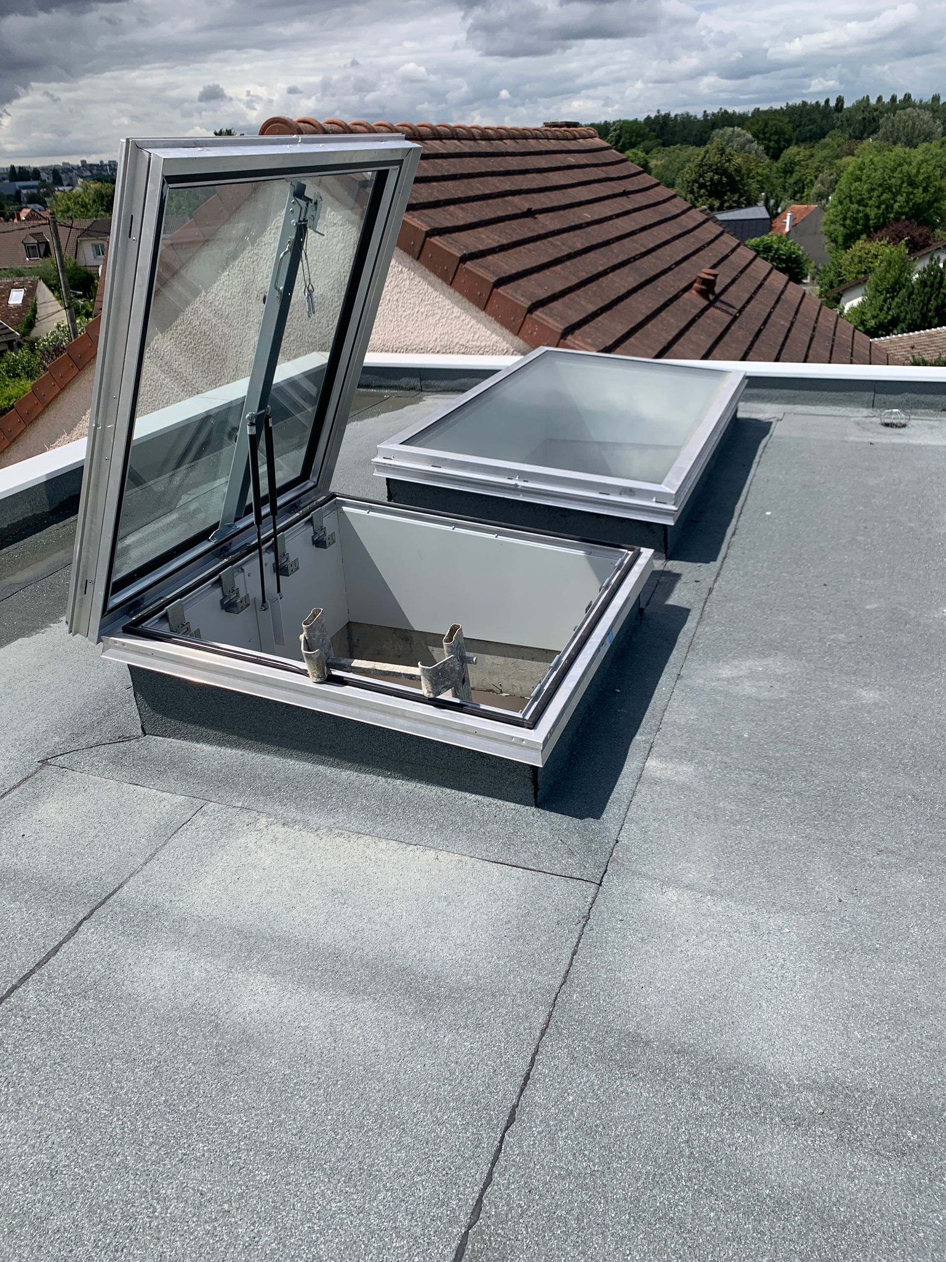 Une trappe de toit ouverte avec un toit vitré sur un toit plat, une deuxième trappe étant fermée. Ciel nuageux.