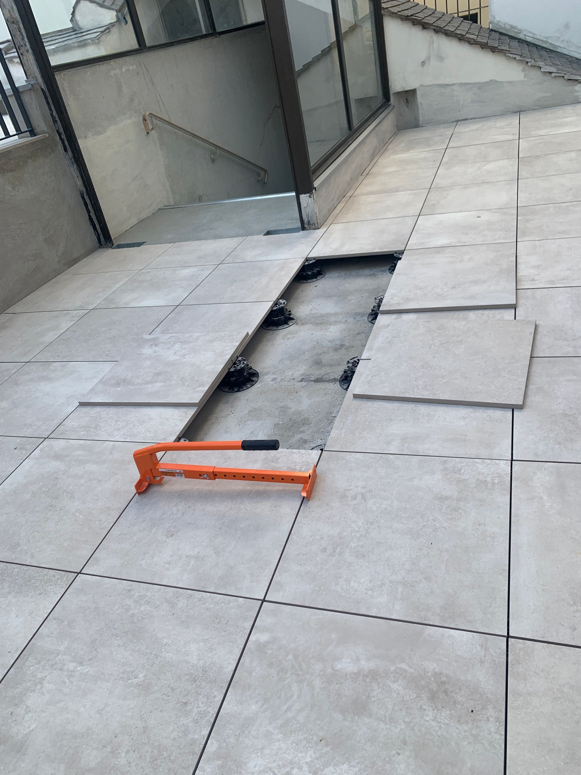 Carrelage en cours d'installation sur une surface extérieure, avec pieds de nivellement et un lève-carreaux orange visibles.