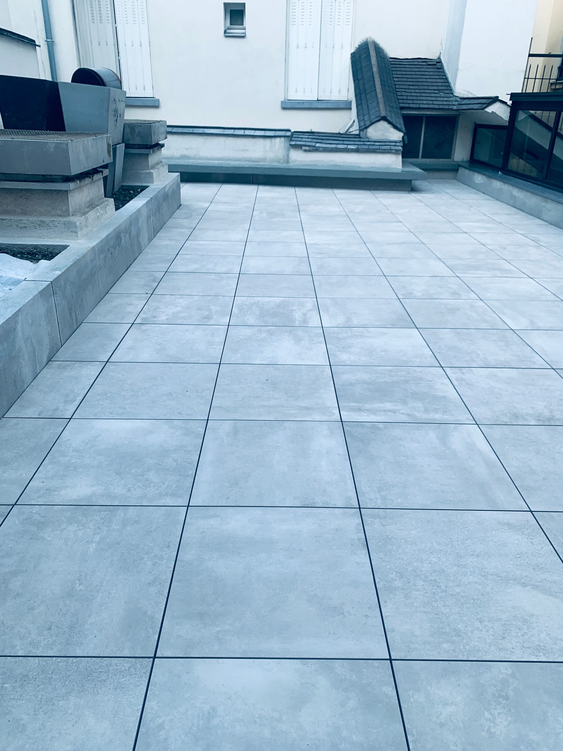 Terrasse carrelée grise avec de longues dalles rectangulaires, adjacente à un muret et des bâtiments en arrière-plan.