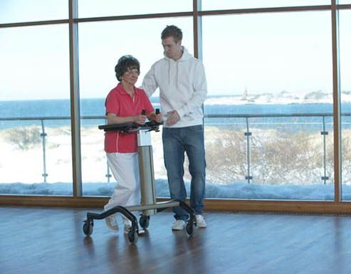 Un hombre ayuda a una mujer con un andador frente a una ventana grande.