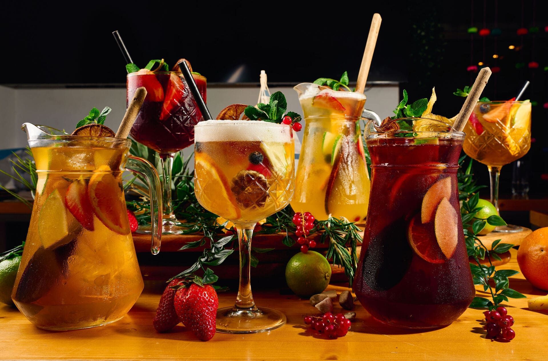 Varias jarras y vasos llenos de sangría colorida, decorados con fruta fresca y hierbas sobre una superficie de madera.