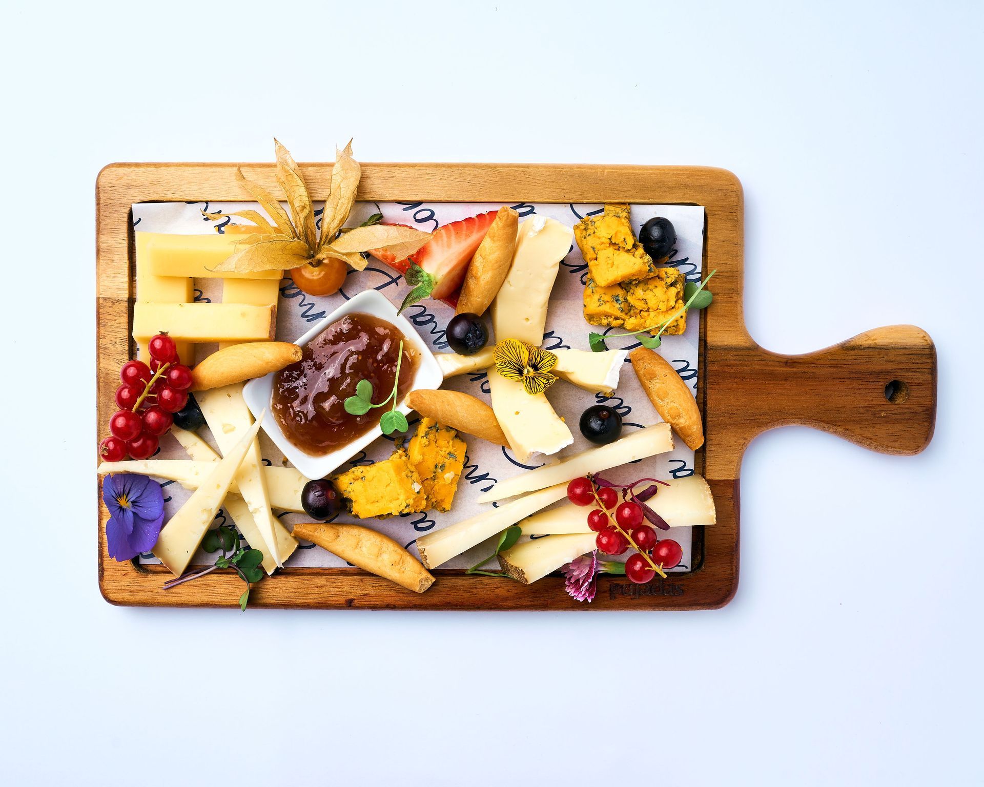 Una tabla de cortar de madera cubierta con queso, fruta y un vaso de mermelada.